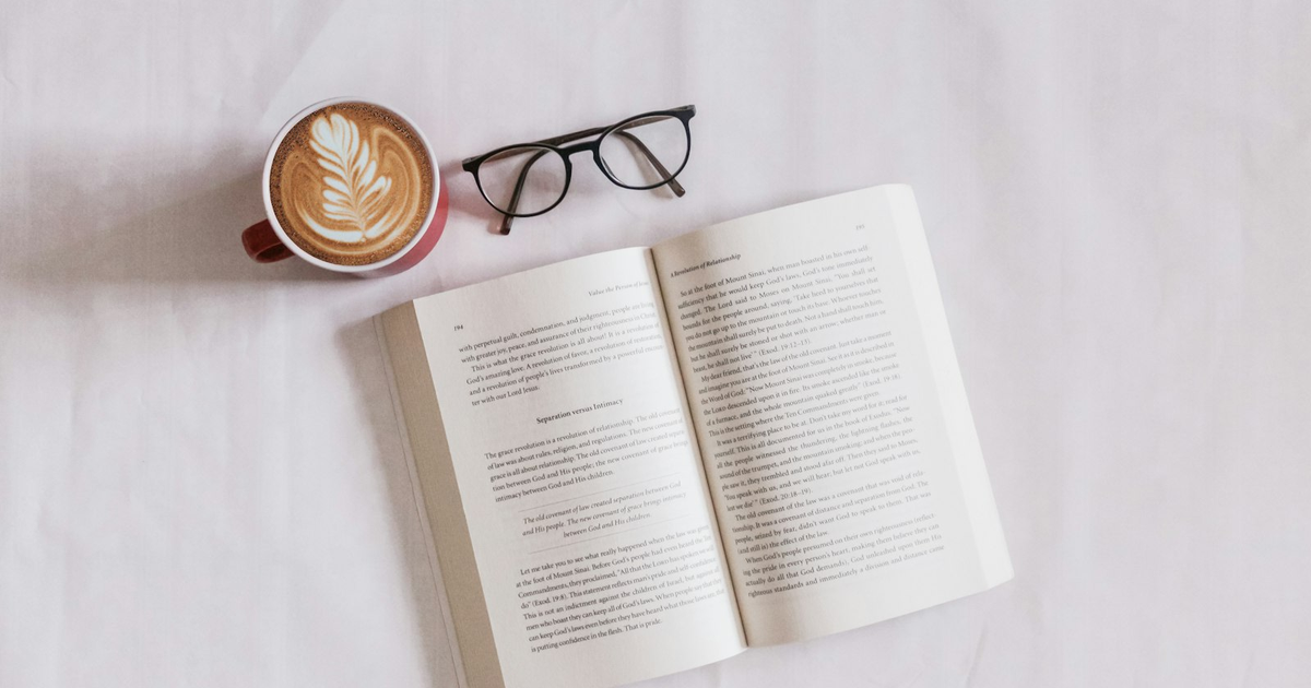 Open book, eyeglasses, and a latte on a white table.